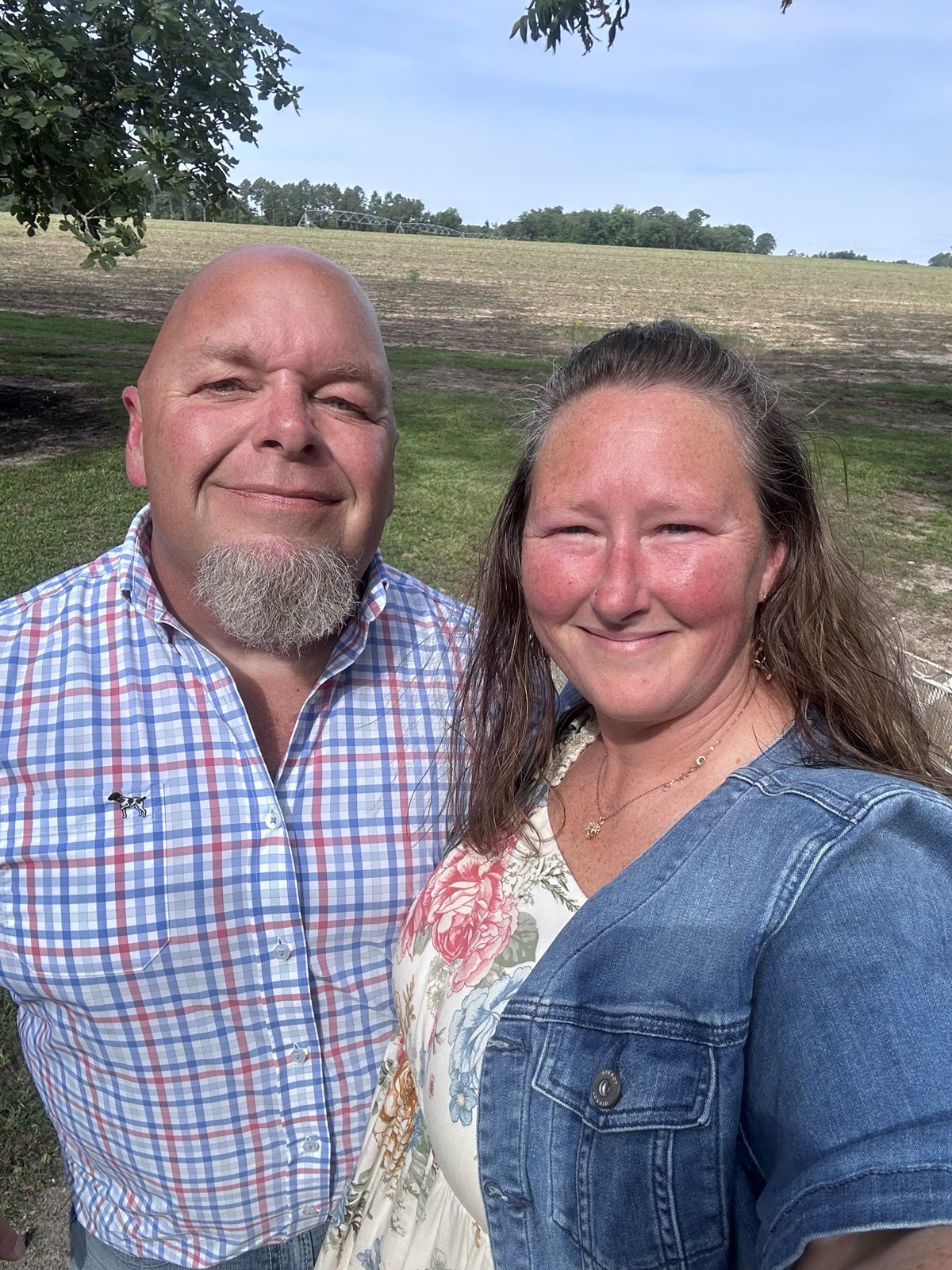Kristie and her husband smiling together outdoors, a farm field behind them