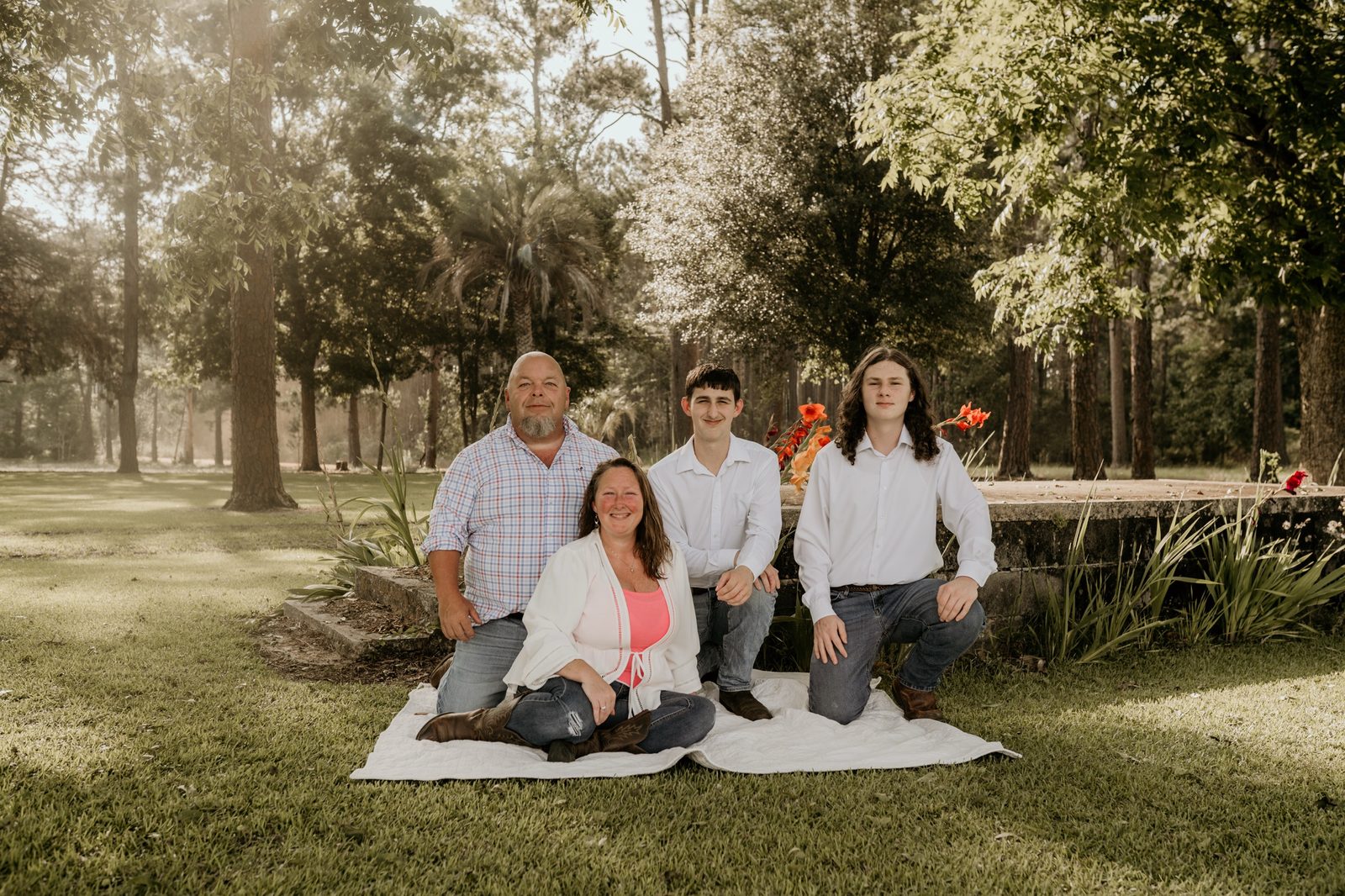 Kristie sitting on a white quilt in the grass with her husband and their two teenage boys kneeling around her, tall pines behind them