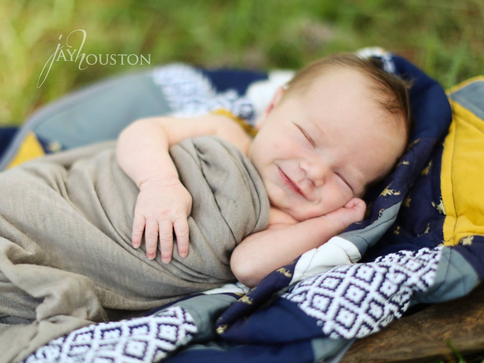 A newborn asleep and smiling on a quilt in the grass